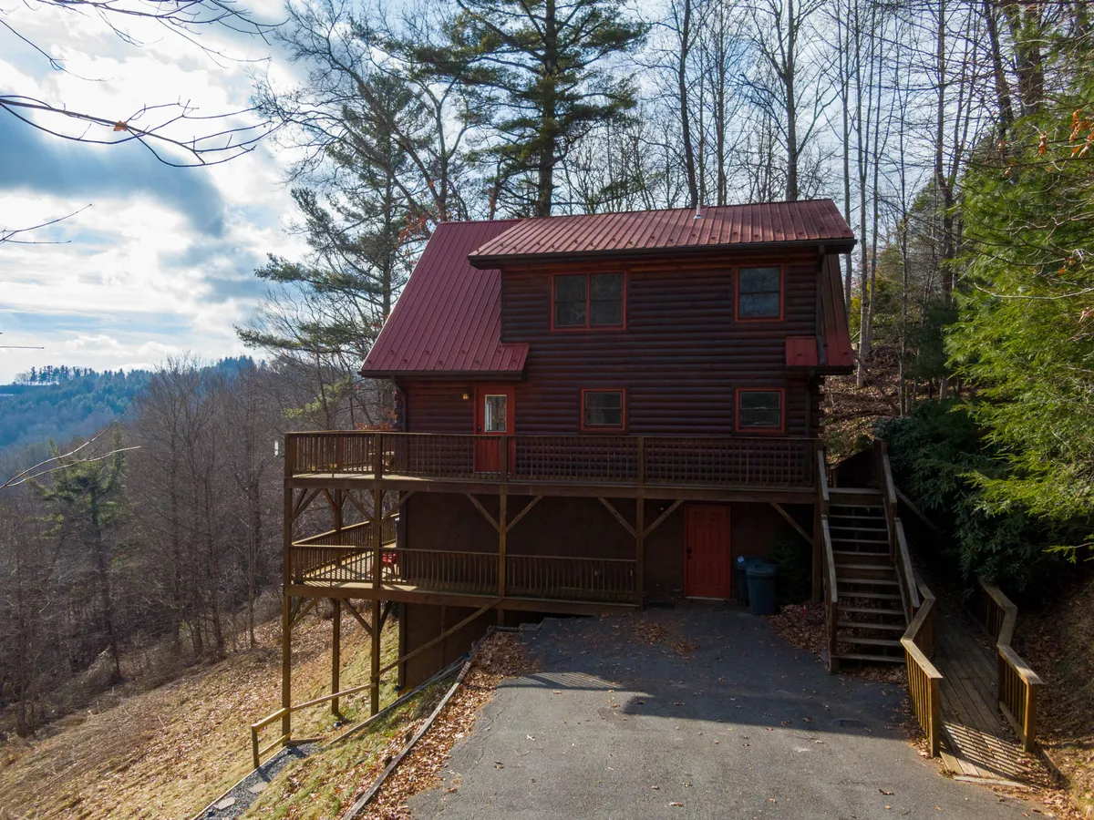 The exterior of a cozy log cabin with a wooden deck, surrounded by trees and featuring a striking red metal roof. The deck invites outdoor relaxation, family gatherings, and observation of the surrounding wilderness.