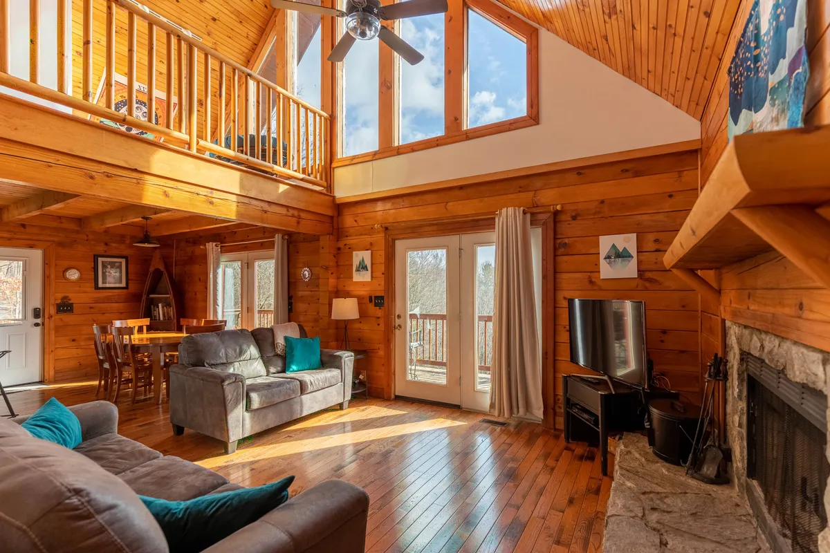 This image shows a sunlit living room featuring large windows, a wooden ceiling, a prominent wood-burning fireplace, and comfortable seating arranged to create a cozy gathering space. The room feels open and is ideal for family relaxation or entertaining guests.