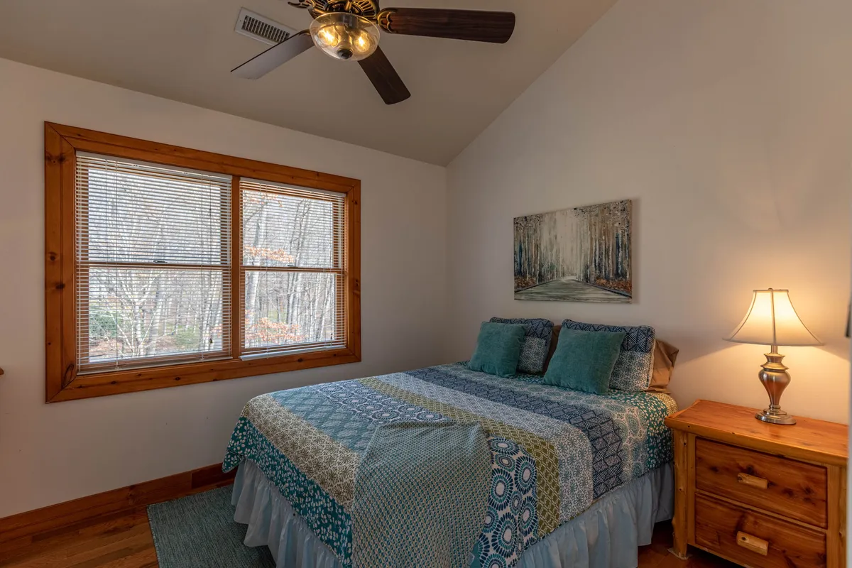 A serene bedroom with a comfortable bed, large windows allowing ample daylight, a classic painting above the headboard, and a soft bedside lamp. The room feels peaceful and private, making it suitable for both relaxation and quiet retreat.