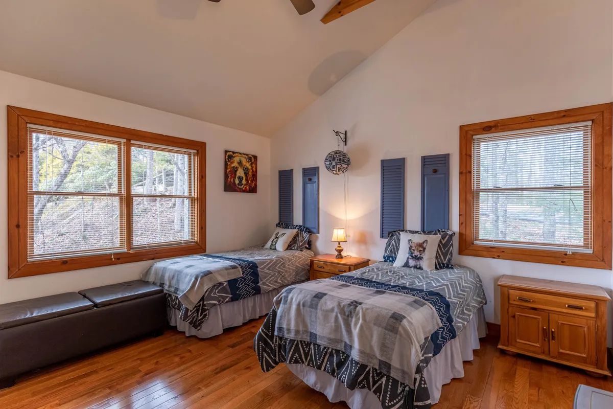 A bright, welcoming bedroom featuring twin beds with crisp linens, large windows letting in natural light, wooden furniture, and tasteful decorative artwork. The setting is airy and ideal for relaxation, reading, or sharing time with siblings or friends.