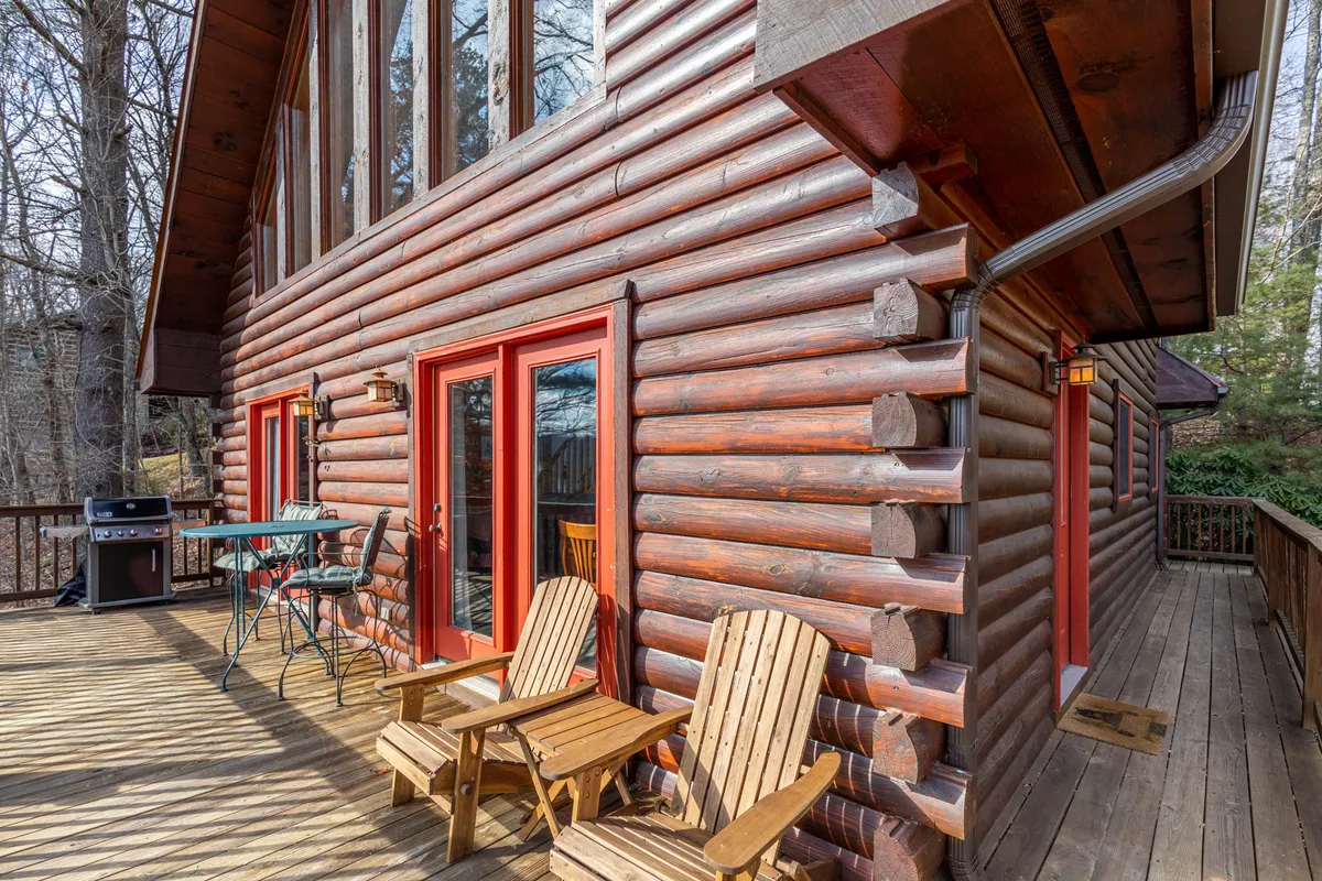 A view of the log cabin exterior with large windows, a wooden deck furnished with Adirondack chairs, and tree-filled surroundings. The scene hints at both accessibility and comfort, with space for outdoor dining and relaxation.