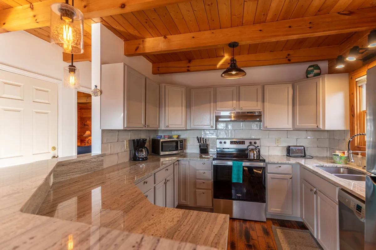 A close-up of the kitchen area, highlighting granite countertops, stainless steel appliances, wooden ceiling, and stylish light fixtures—offering a modern yet rustic feel.