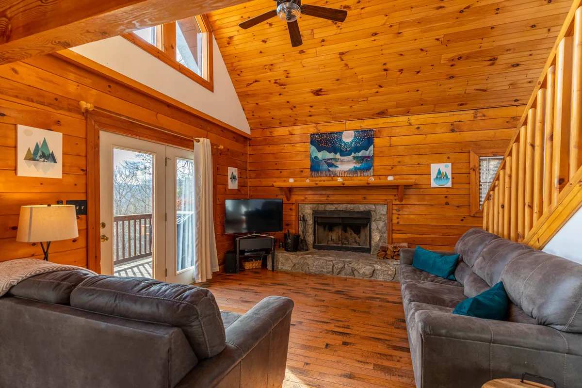 A spacious living area featuring a wooden ceiling, a large fireplace as the centerpiece, expansive windows letting in natural light, and a comfortable sofa arranged for socializing. Art adorns the walls, and the warm ambiance is perfect for relaxing after a day outdoors.