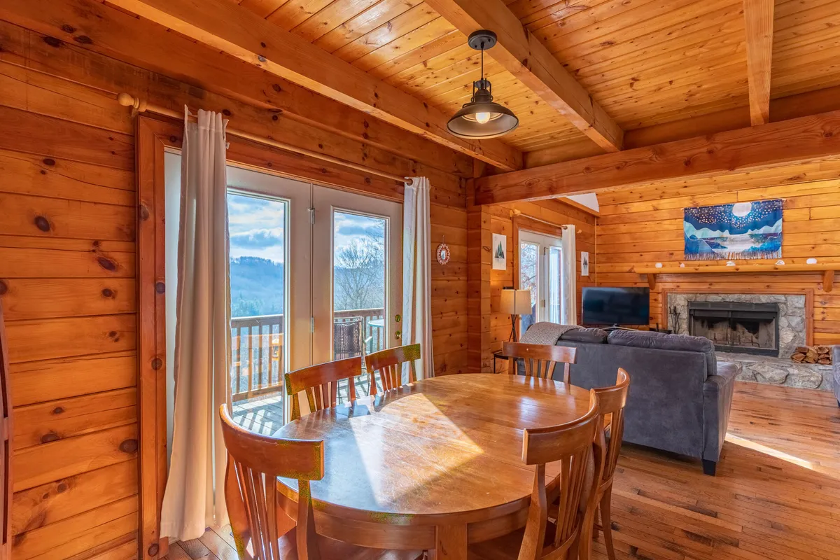 A bright, airy interior space with a large dining table set near expansive windows, a cozy fireplace, and a comfortable sofa. The windows frame scenes of the outdoors, visually blending indoor and outdoor experiences. Warm natural light and mountain artwork create a sense of being surrounded by nature even while inside.