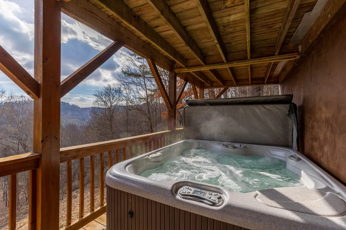 This inviting exterior shot features a relaxing hot tub on a wooden deck with open beams, positioned to overlook expansive mountain views. The image captures the transition between indoor comfort and the wild outdoors, offering a prime spot for soaking in natural beauty, watching sunsets, or stargazing.