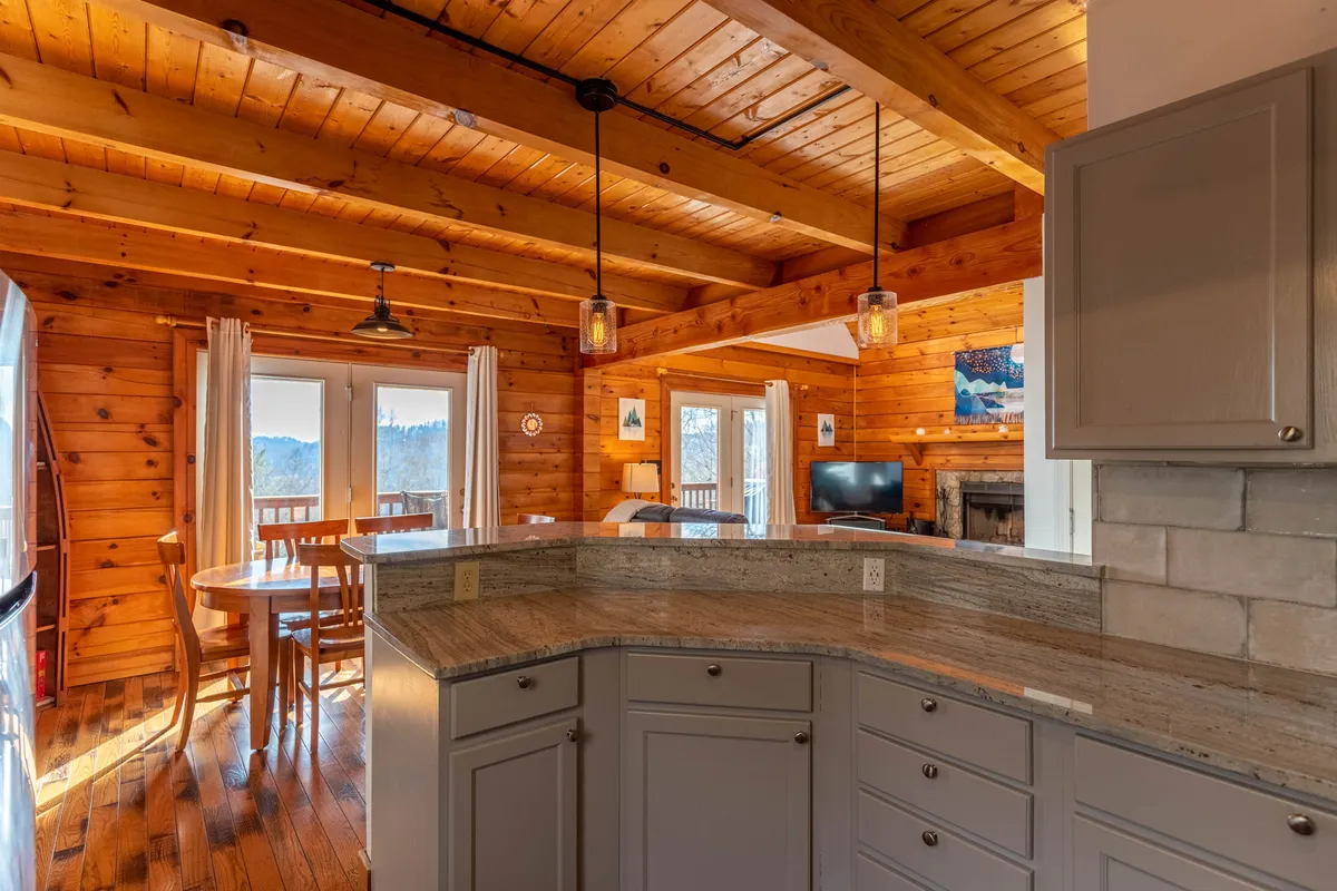 A spacious, open-plan kitchen and dining area with a granite kitchen island, a dining table for six, large windows, wooden beams, and a fireplace visible in the background. The layout flows naturally from cooking to dining and relaxation.