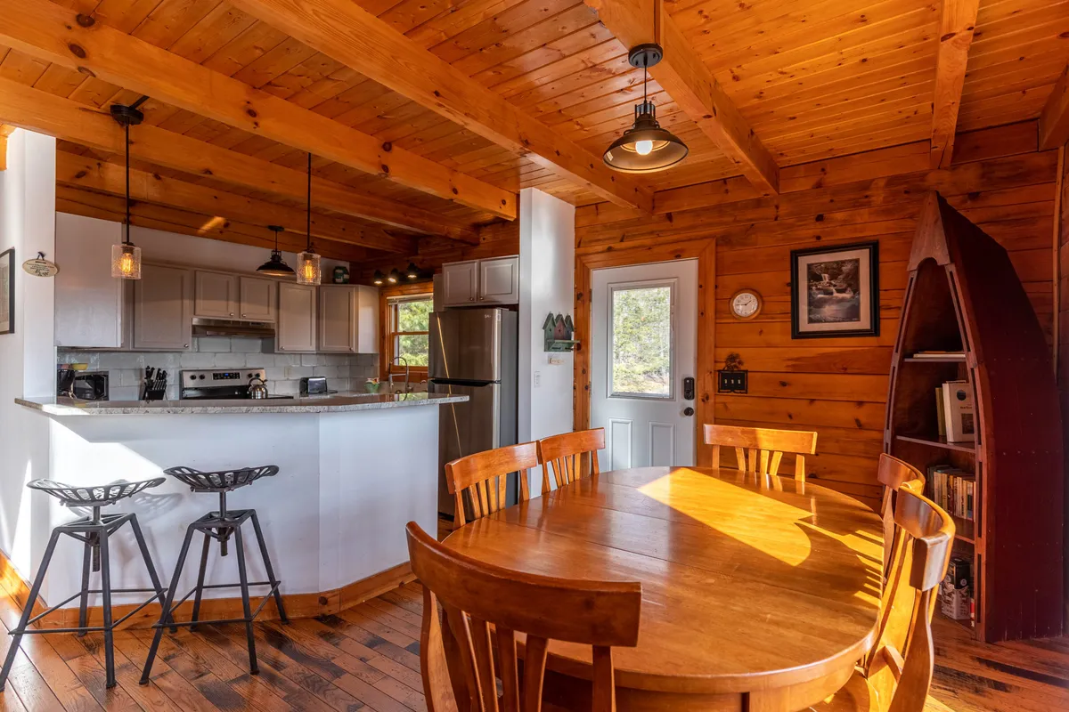 This image captures a sunlit dining area with a wooden table, bar stools, and an open door leading outside. The open layout allows for easy movement between inside and out, and the visible bookshelf and kitchen countertop suggest spaces for gathering, entertaining, and seasonal activities.