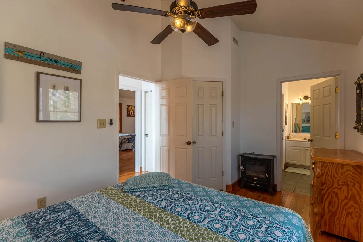 A cozy bedroom is shown with a quilted bedspread, a ceiling fan, and open doorways leading to other rooms. The space is well-lit and inviting, reflecting a comfortable environment suitable for guests seeking accessible accommodations.