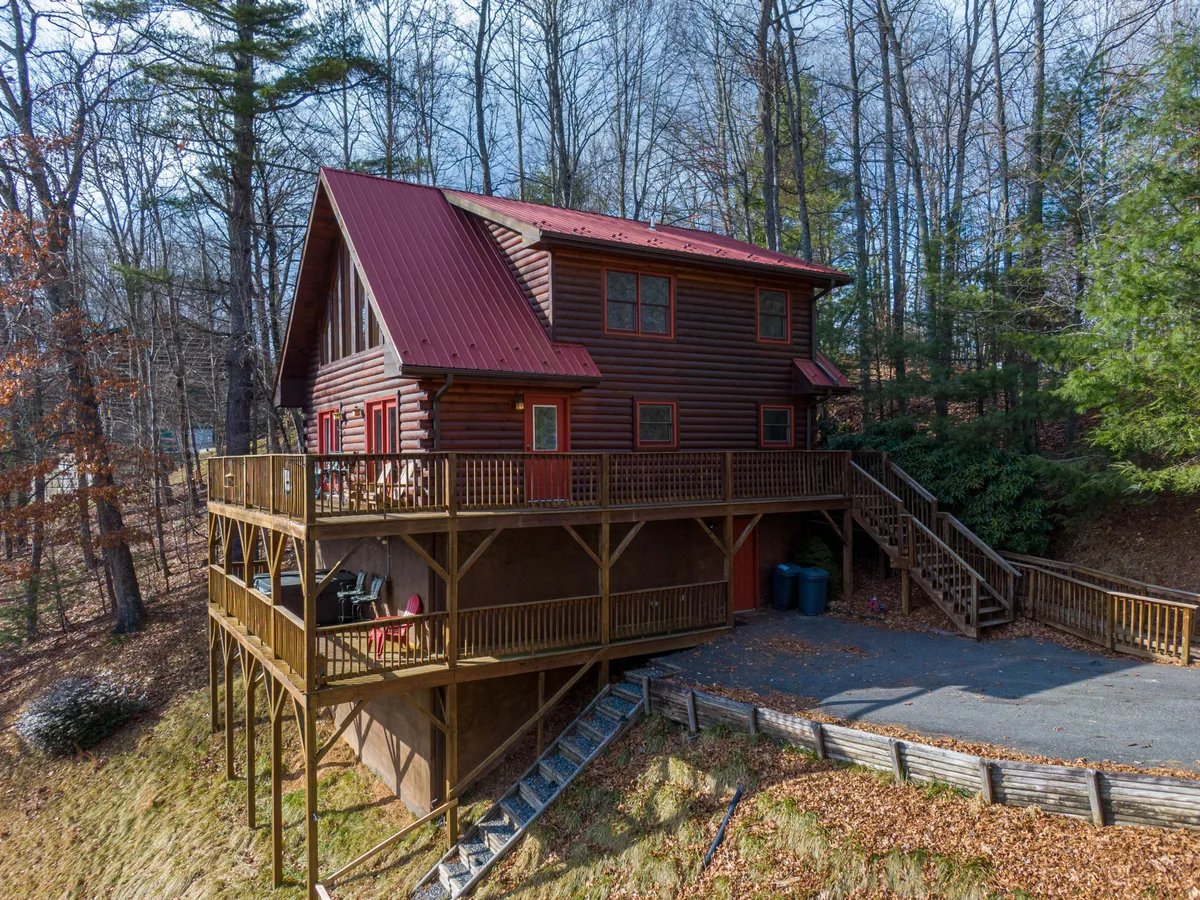 This image shows the exterior of the log cabin, highlighting the wooden deck, red metal roof, and surrounding trees. The deck offers ample space for outdoor gatherings, relaxation, and enjoying nature. The log cabin structure sits harmoniously in a tranquil, wooded setting, evoking a sense of retreat and adventure.