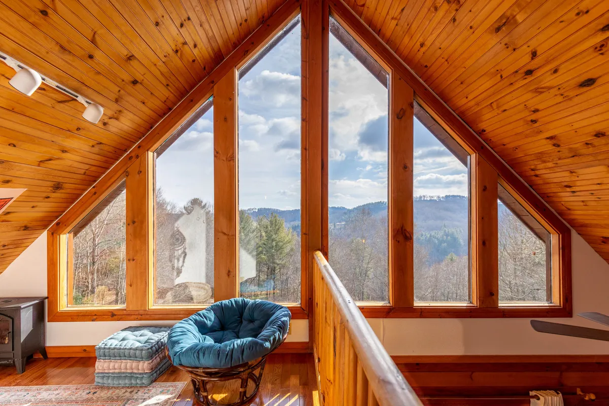 An open loft space with dramatic triangular windows, rustic wooden beams, and a plush blue chair. Natural light floods the area, and the windows offer stunning mountain views, creating a tranquil spot for morning coffee, meditation, or simply enjoying the scenery.