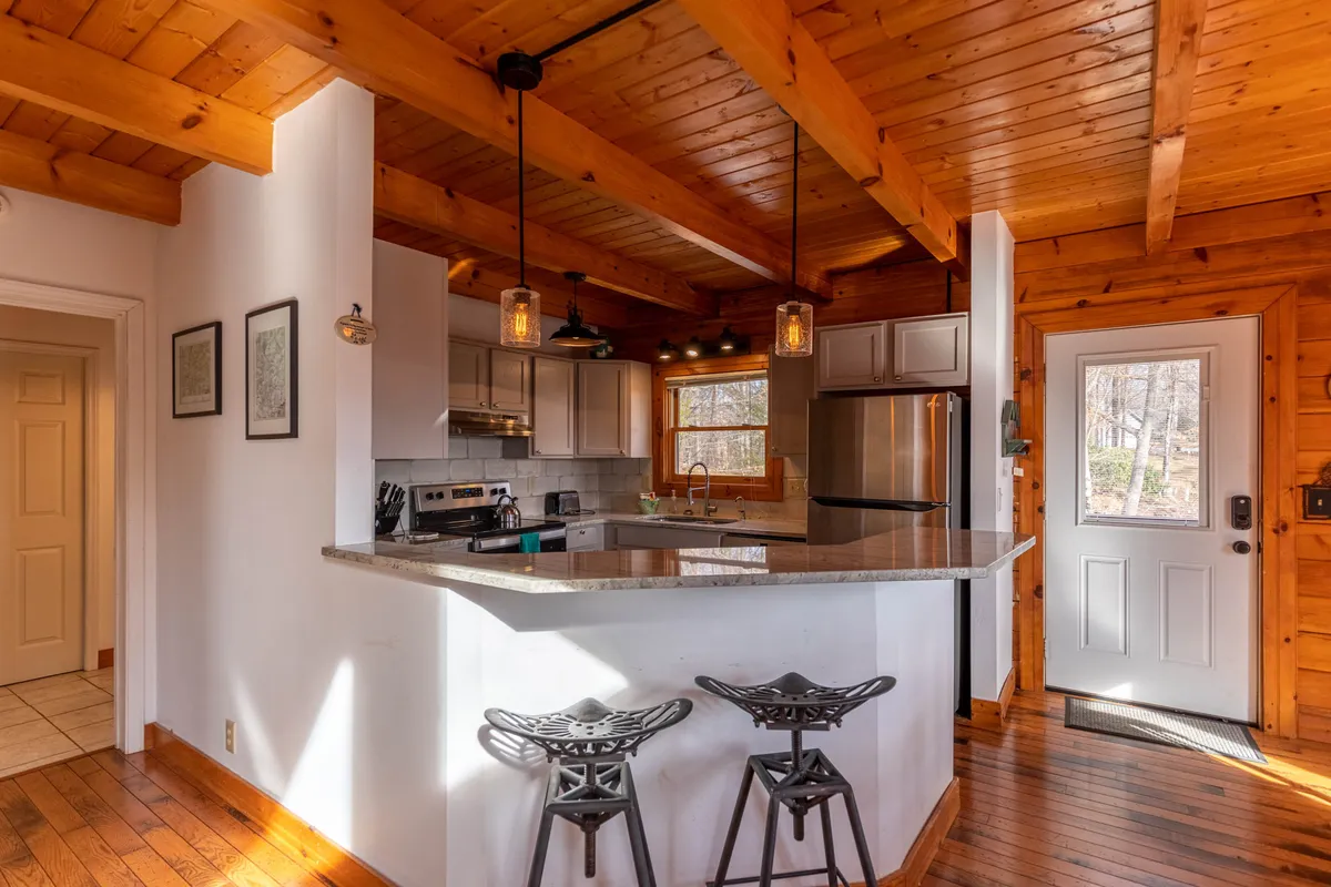 A bright, welcoming kitchen space with wooden beams, granite countertops, stainless steel appliances, and cozy bar stools. Sunlight streams in, illuminating the area where guests might savor a quiet morning coffee or prepare a meal together.