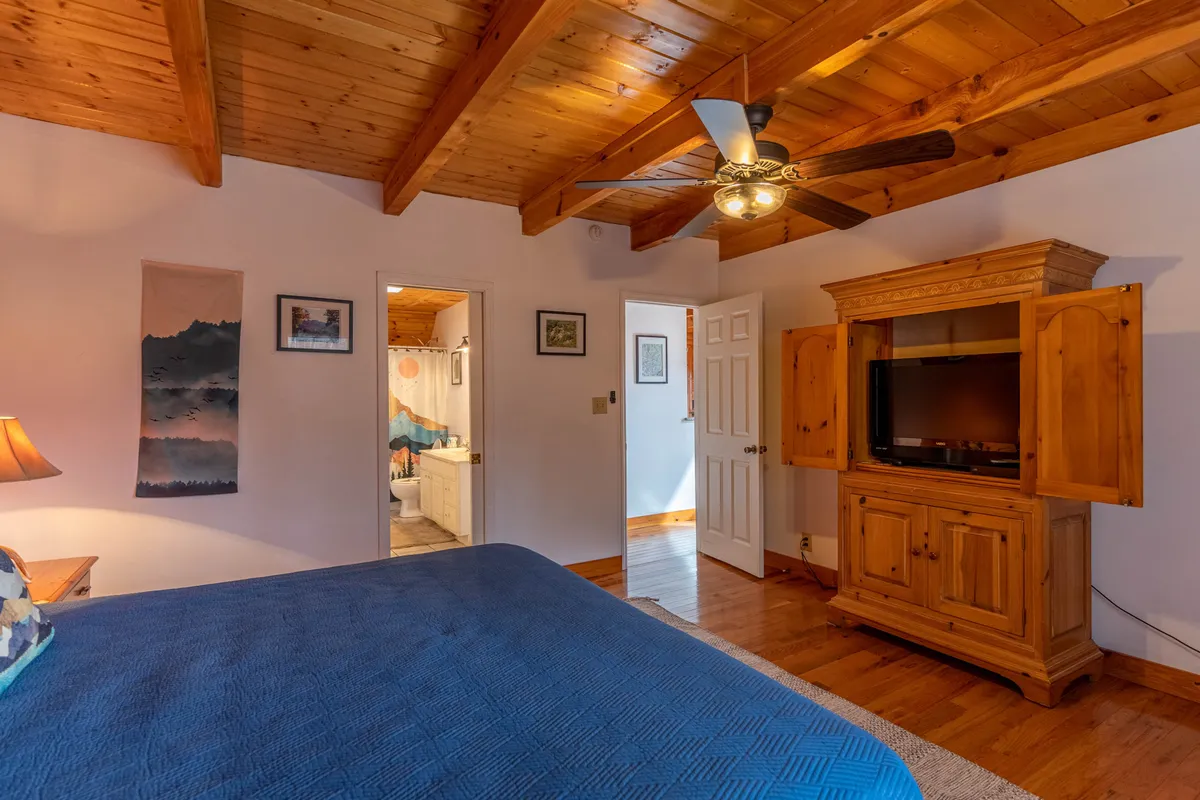 This image presents a cozy bedroom with wooden ceiling beams, a blue bedspread on a comfortable bed, a TV cabinet, and a doorway leading to an adjacent bathroom. The room feels open and accessible, with calming decor and a soft, inviting color palette. The layout suggests ease of movement, supporting both comfort and accessibility.